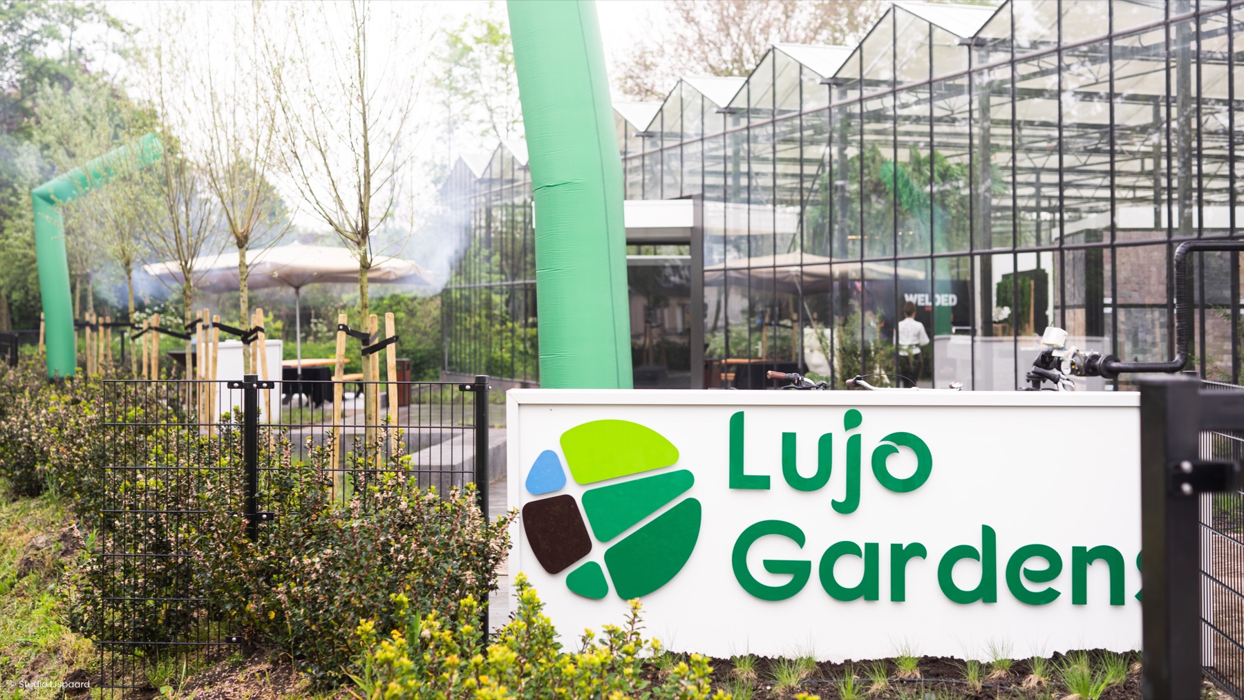 Entree van LujoGardens in Uden, met het naambord, de glazen showroom en groen langs het pad.